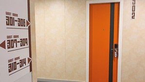 Un pasillo con indicaciones para habitaciones de huéspedes, una puerta con una franja negra y un letrero con número fijado a la pared.