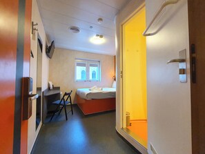 A narrow hallway with a bed, a desk, and a chair. There is a window with a view of a building. - TL Hotel Budapest (Budapest)
