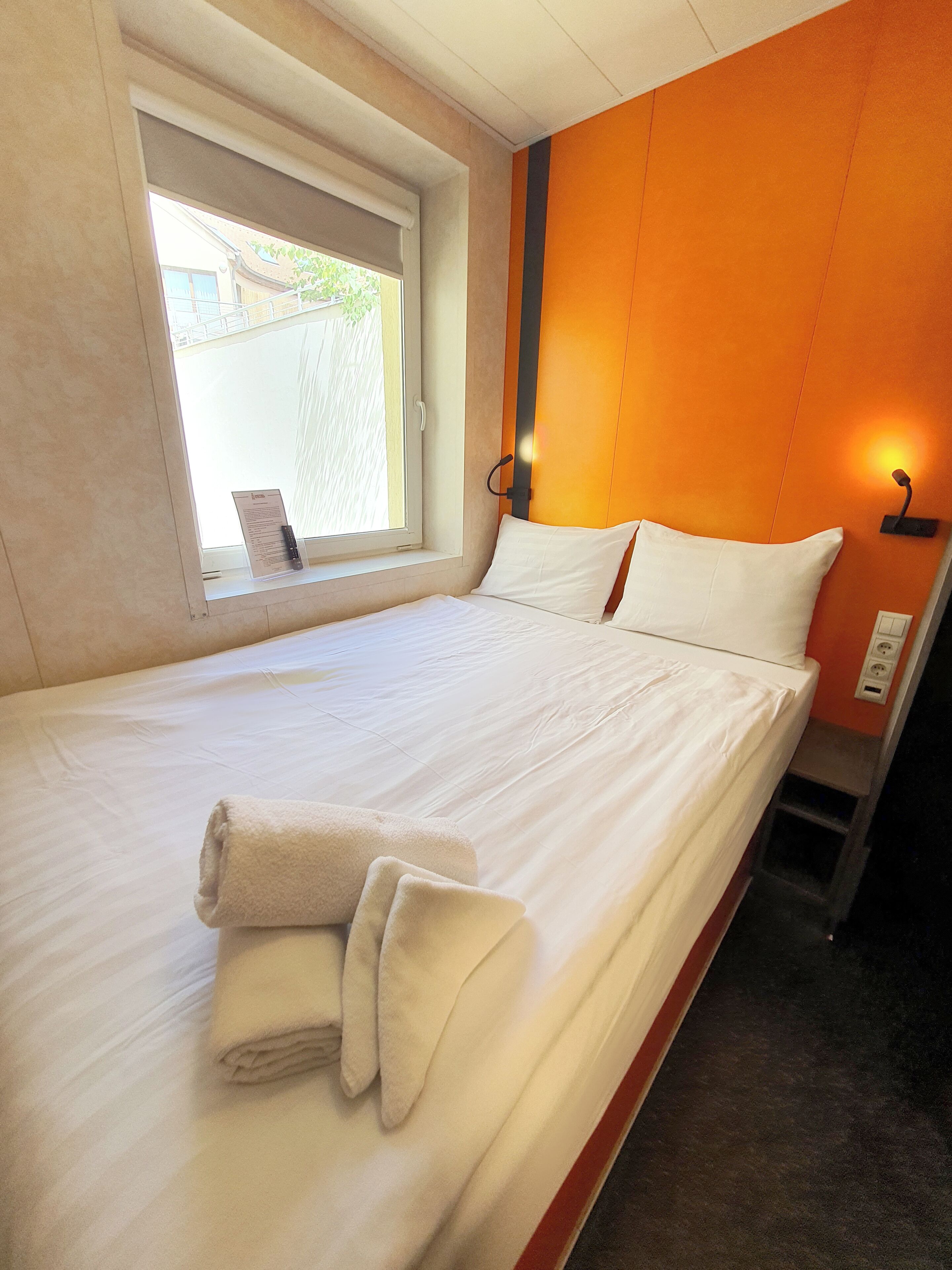 A hotel room with a bed, a window, and a wall-mounted light fixture.