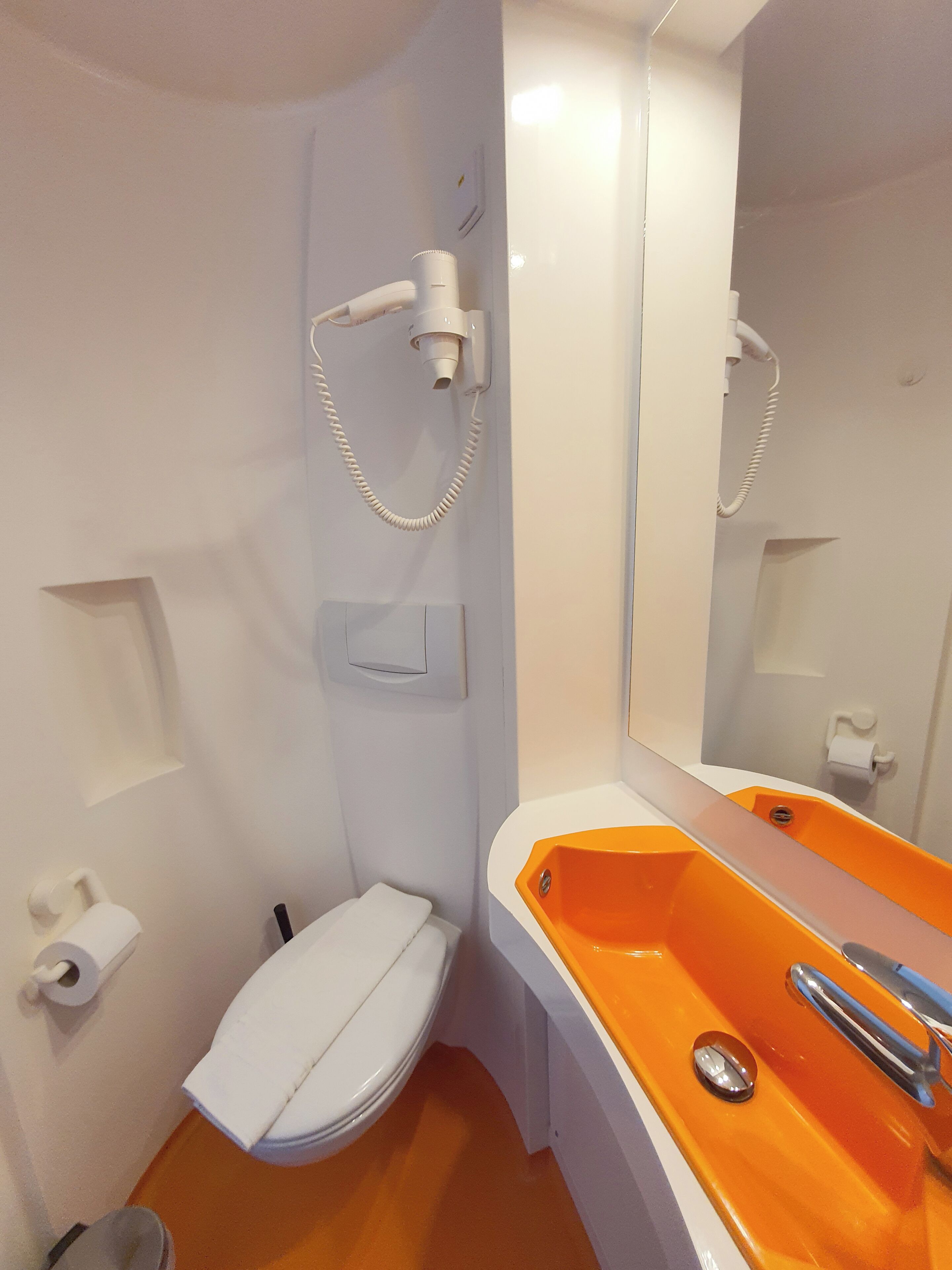 A bathroom with a white toilet, a white sink with an orange basin, a handheld hair dryer mounted on the wall, and a mirror reflecting the bathroom.