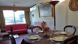 Een eetruimte met een ronde tafel voor vier personen, een rode bank, uitzicht over de stad door grote ramen en een schilderij aan de muur.