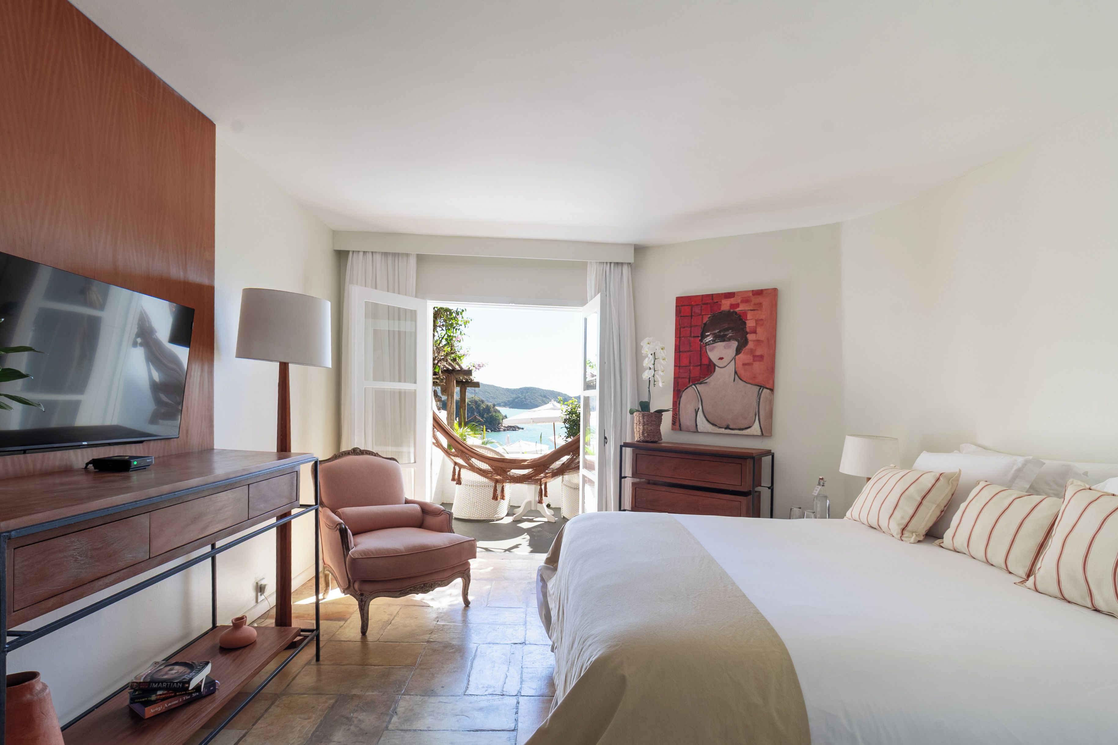 Una habitación de hotel con cama, escritorio con televisor, silla, hamaca y un cuadro en la pared.