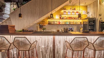 Zona de bar con techo de madera inclinado, una variedad de botellas, vasos y un refrigerador. Hay sillas de mimbre frente a la barra.