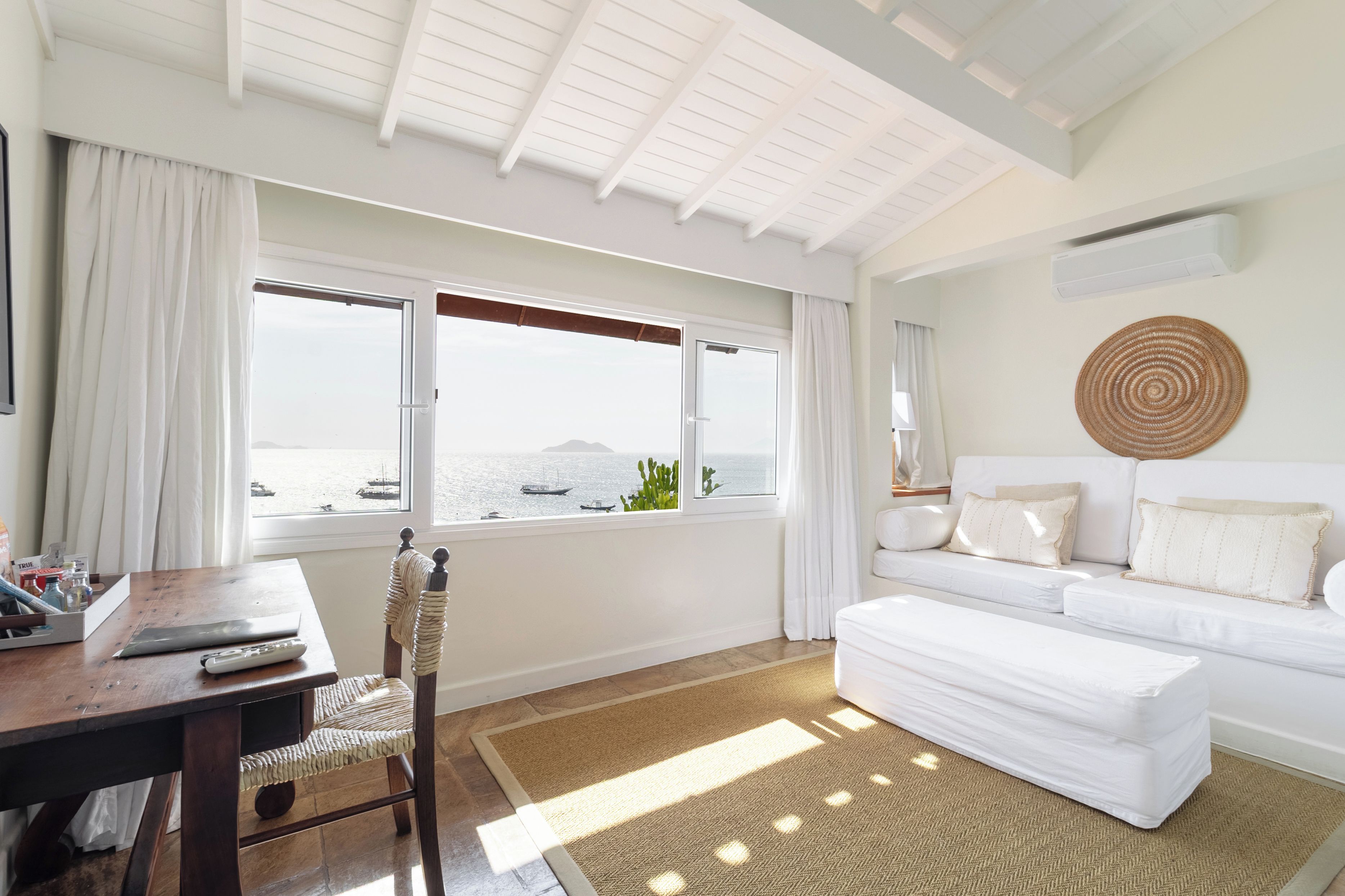 Una habitación de hotel moderna con un ventanal grande, un sofá blanco, una mesa de comedor de madera y una alfombra tejida.