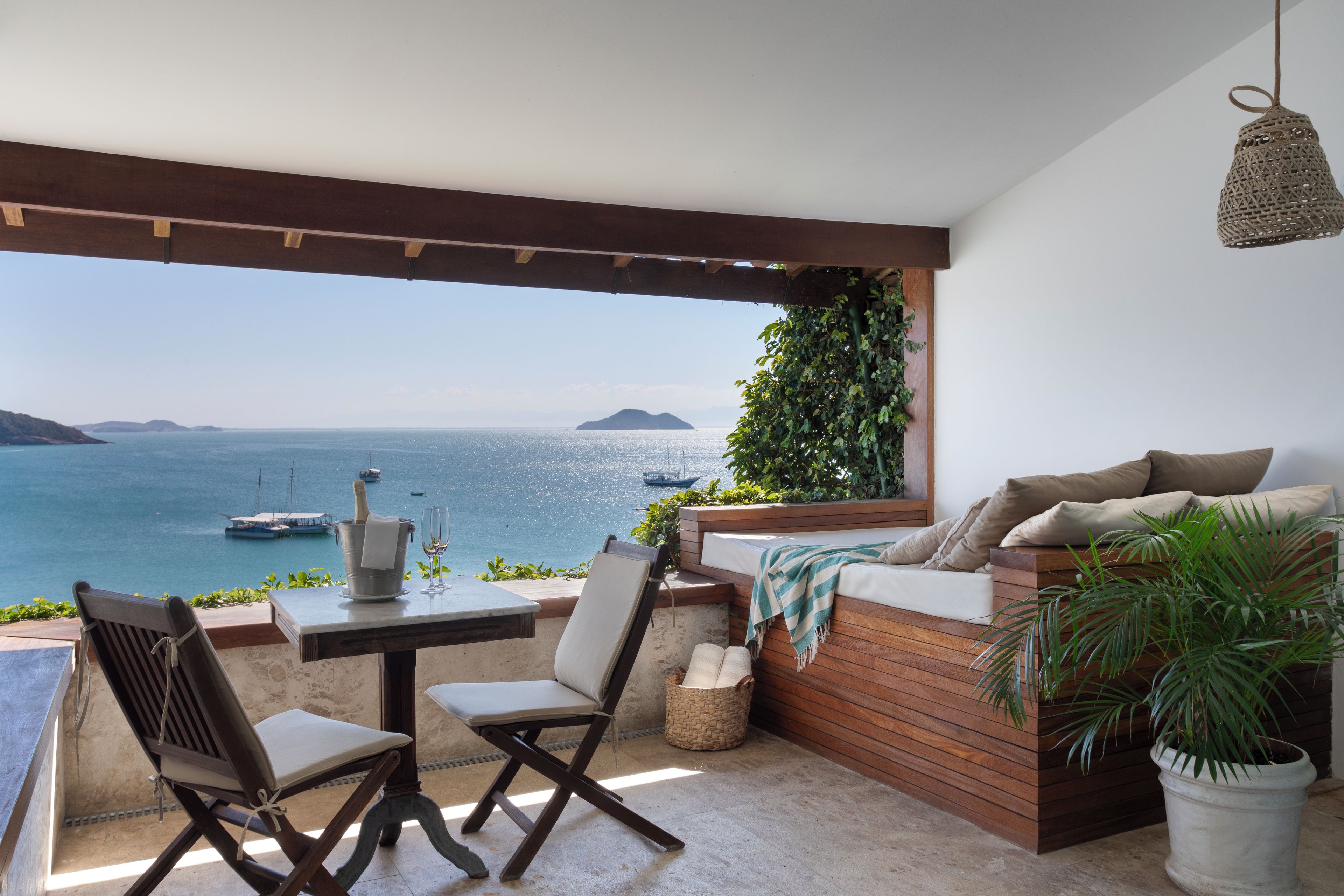 A terrace with a view of the sea, a table with a bottle and glasses, a cushioned seating area, and a hanging light fixture.
