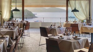 Un restaurante con mesas dispuestas para comer, amplios ventanales con cortinas y vistas al mar y a las montañas.
