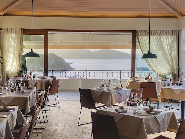 Un restaurante con mesas dispuestas para comer, amplios ventanales con cortinas y vistas al mar y a las montañas.