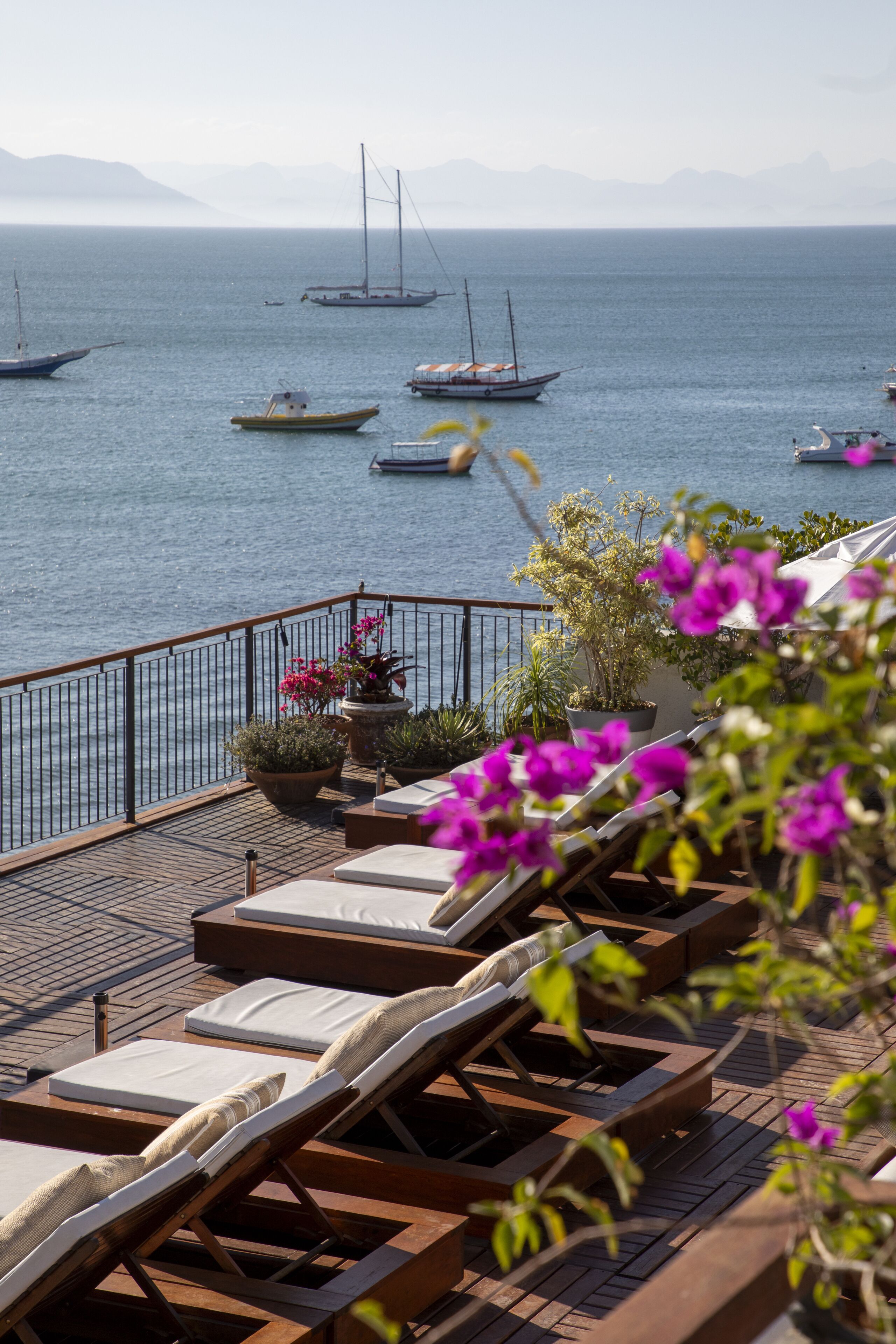 A deck with lounge chairs and potted plants overlooking a calm sea with boats.