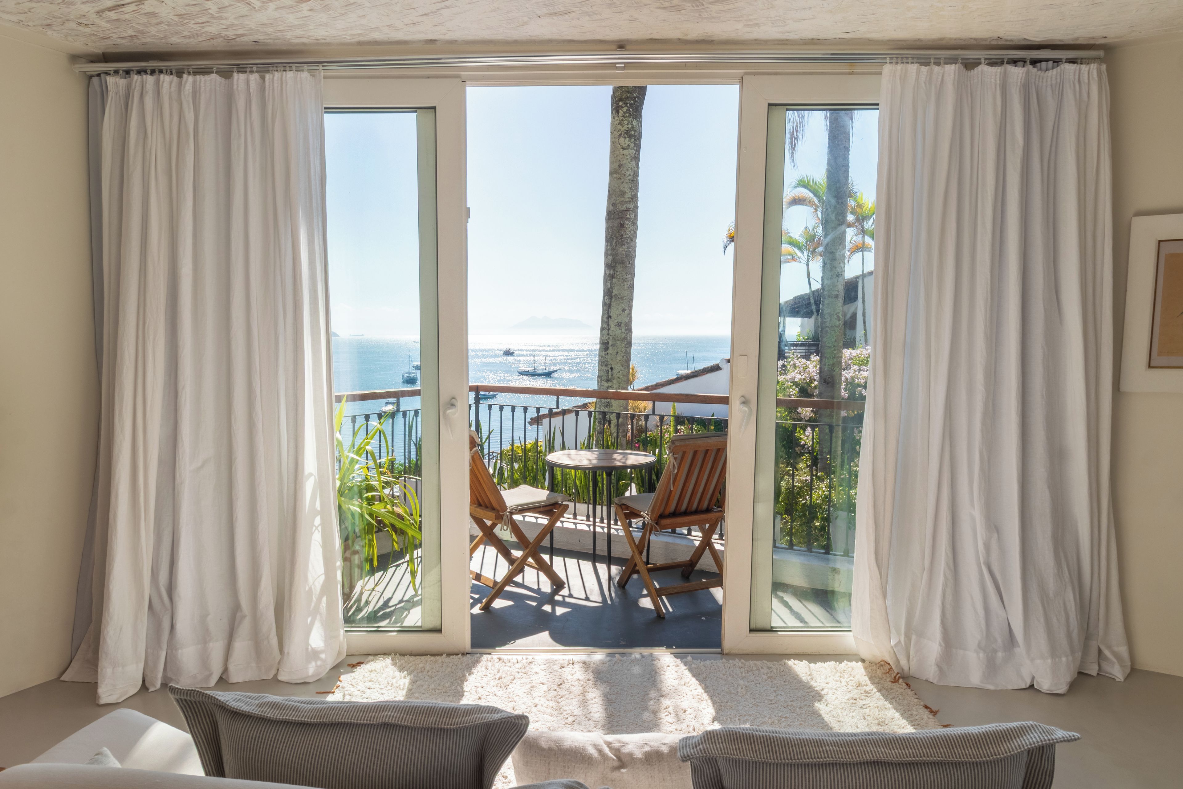 A room with a view of the sea, a balcony with chairs and a table, and a large window with curtains.