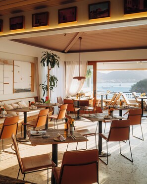 A spacious dining area with multiple tables and chairs, large windows offering a view of the sea, and a ceiling with exposed beams and pendant lighting. - Casas Brancas Boutique Hotel & Spa (Búzios)