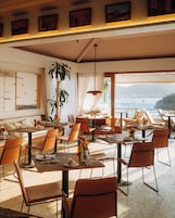 Un comedor amplio con múltiples mesas y sillas, ventanas grandes que ofrecen vistas al mar, y un techo con vigas a la vista e iluminación colgante.