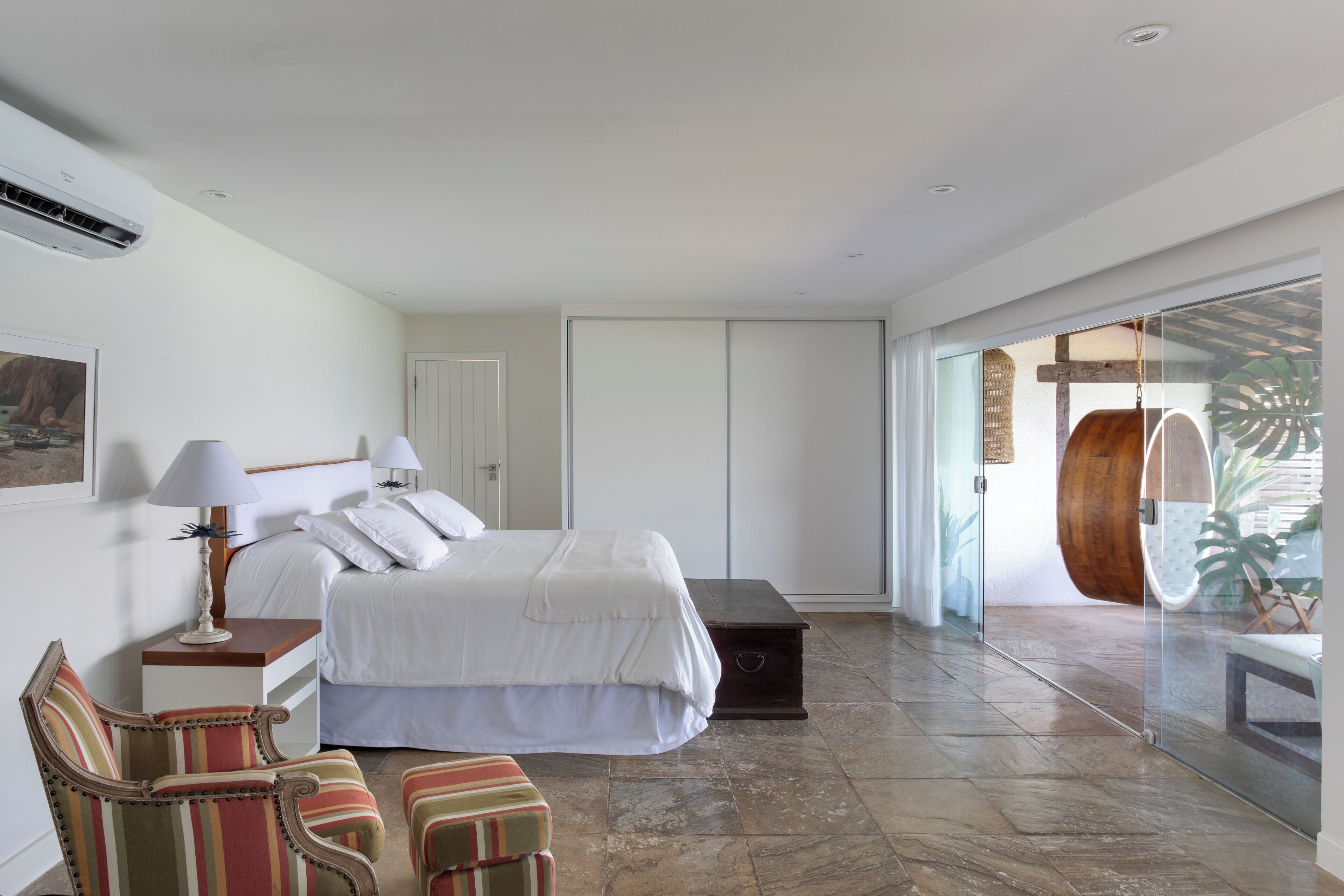 A bedroom with a bed, bedside tables, a chair, a bench, and a glass door leading to a balcony with a wooden barrel and tropical plants.