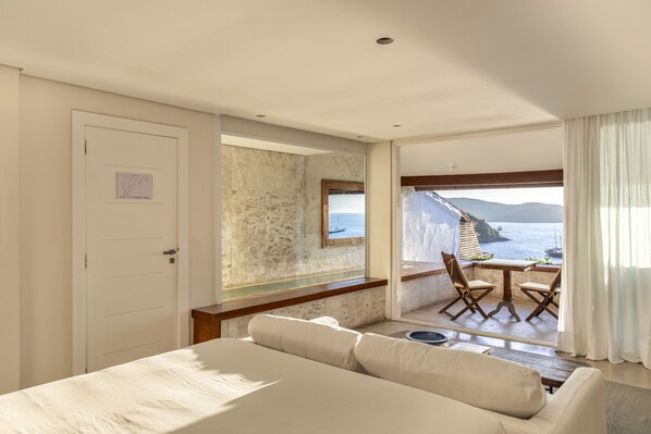 Habitación de hotel con cama, zona de estar con mesa y sillas, vista al mar y una puerta con un letrero.