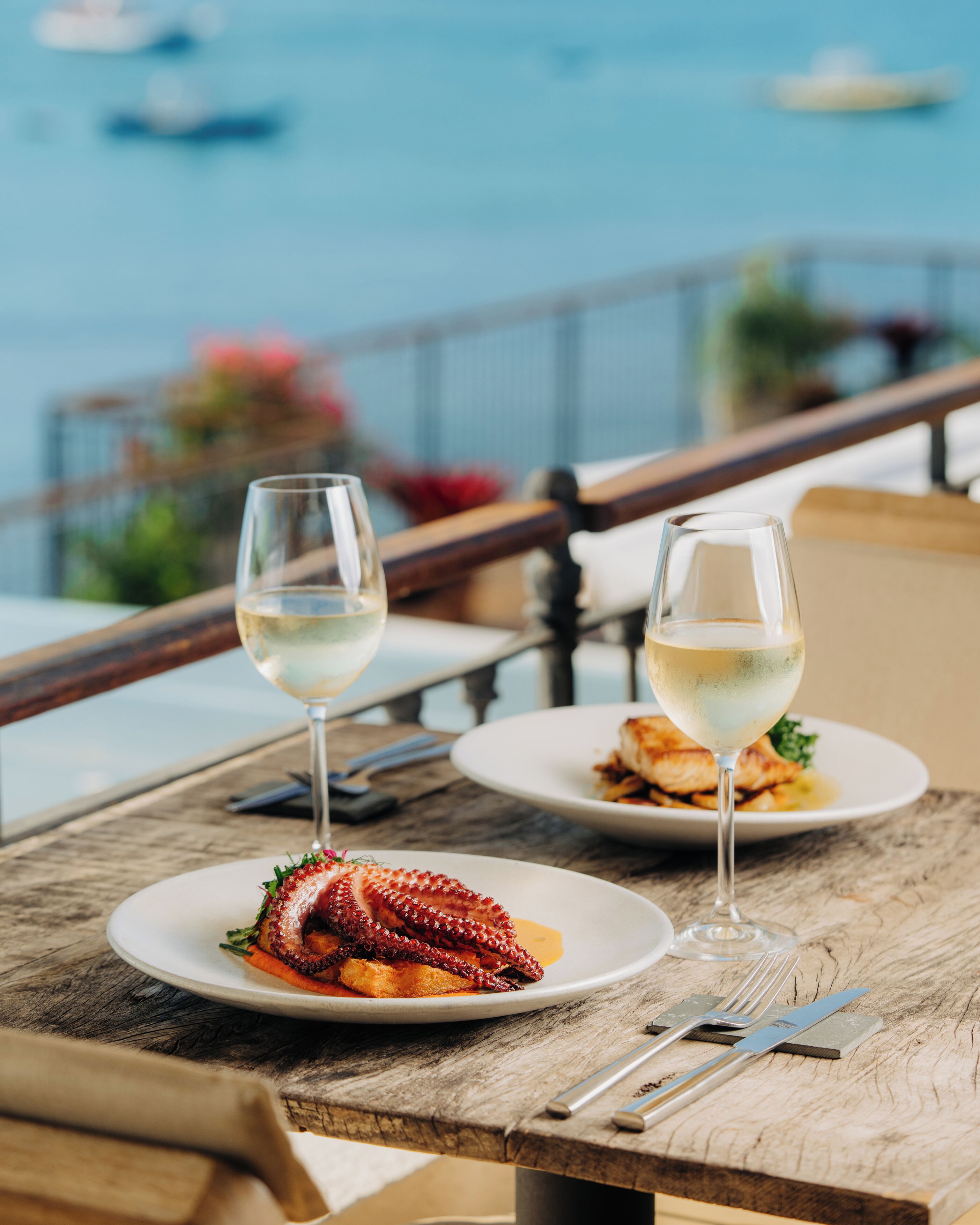 A dining table with two plates of food and two glasses of white wine, set against a backdrop of a scenic view.
