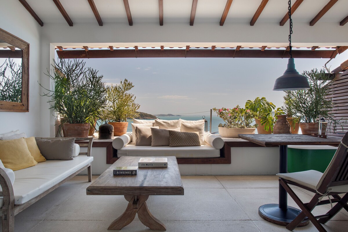 A patio with a sofa, a table, and chairs, surrounded by potted plants and a view of the sea.