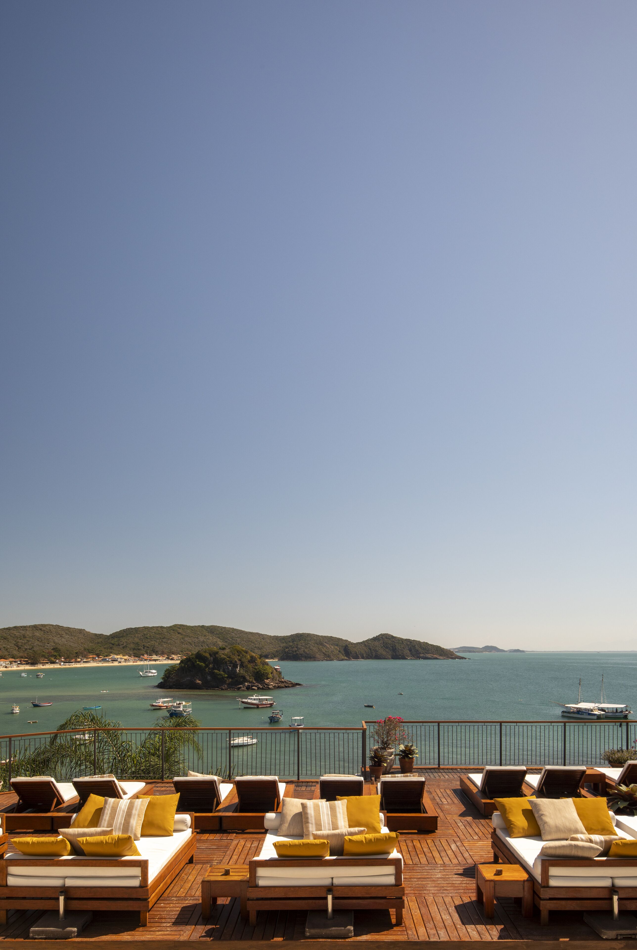 Terraza de madera con sillones y cojines, con vistas a una zona costera con barcos y cielo despejado.