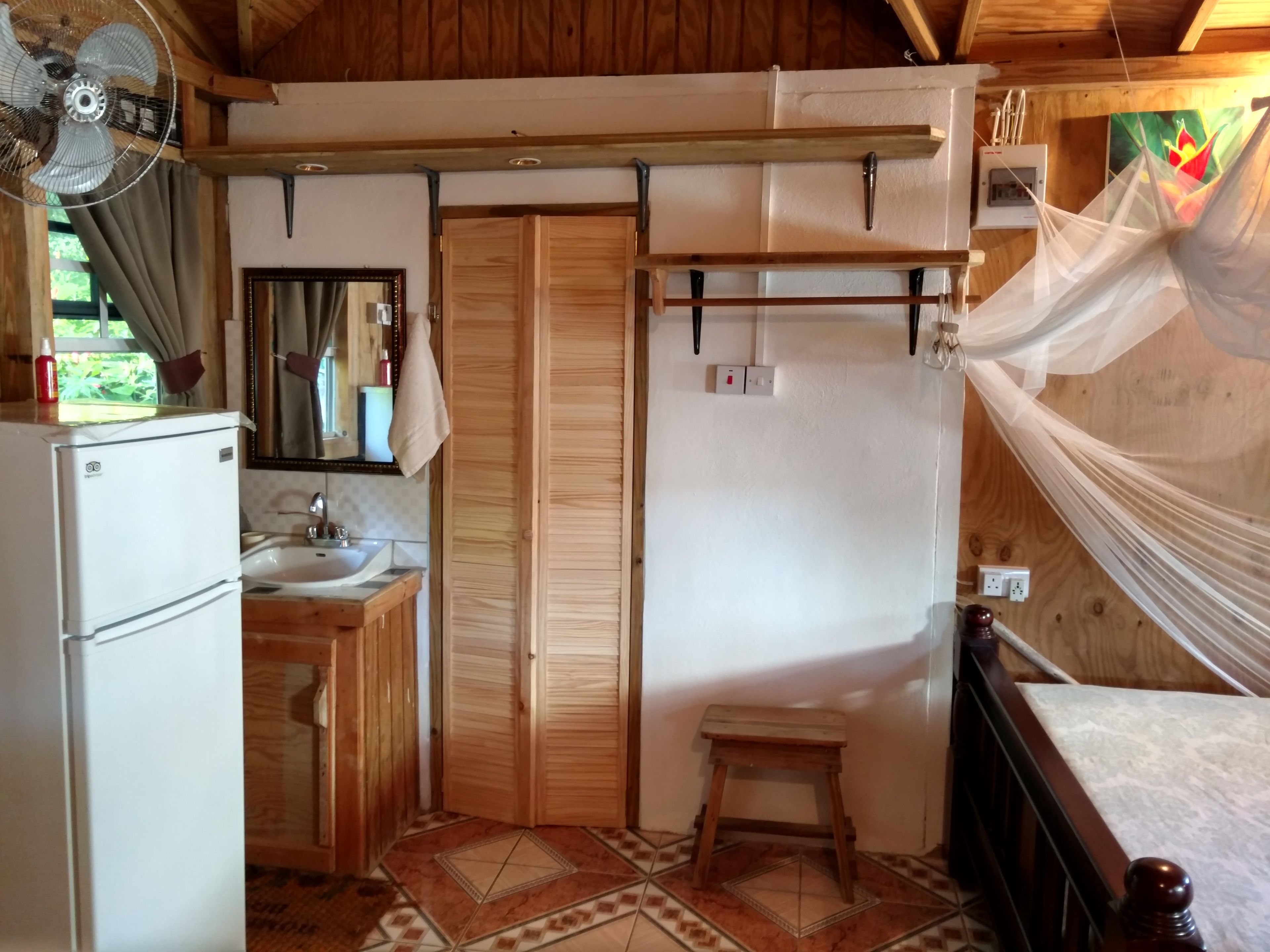 Een kleine kamer met een wastafel, spiegel, houten kastjes, een ventilator en een bed met klamboe.