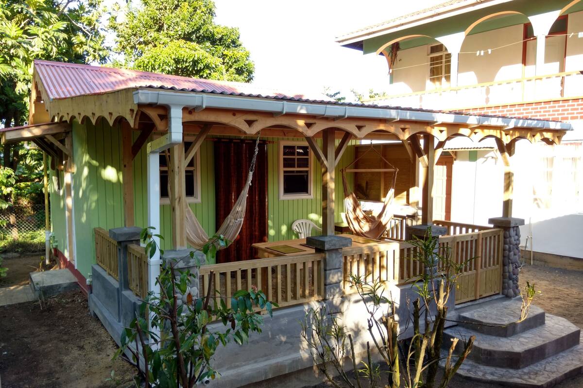A green house with a porch, a hammock, and a small outdoor seating area.