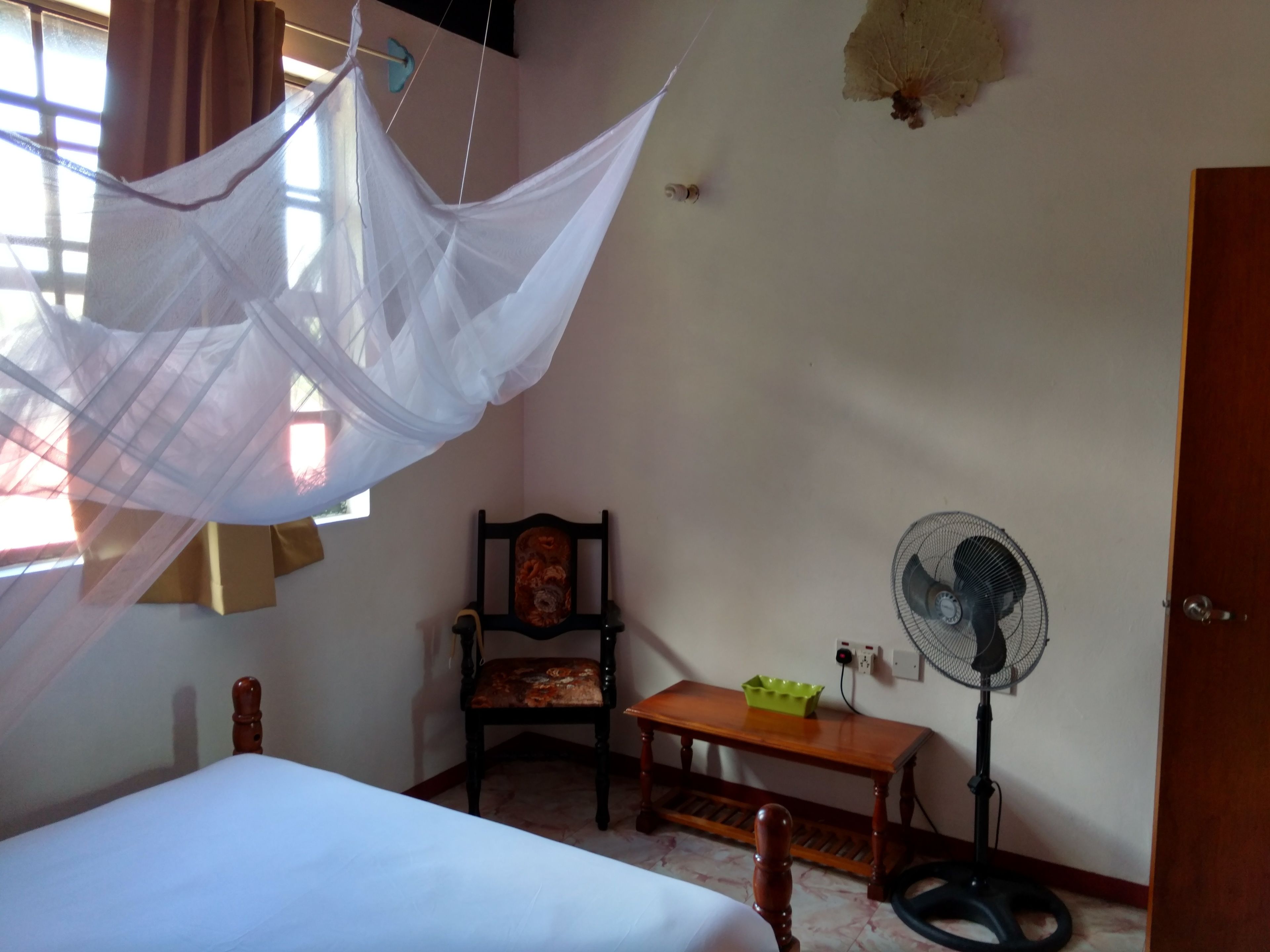 Een slaapkamer met een bed, een stoel, een nachtkastje, een staande ventilator en een klamboe.