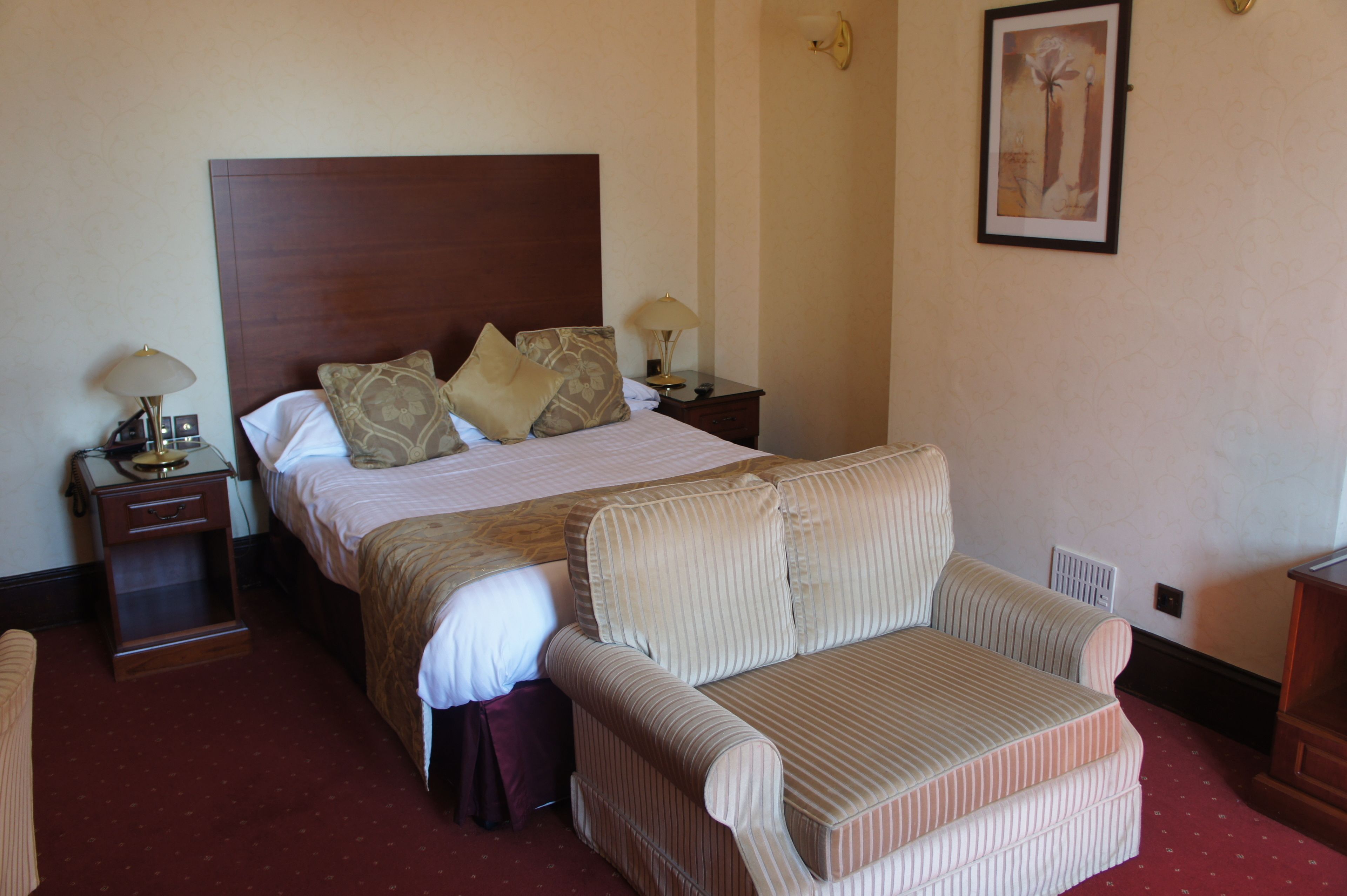 A hotel room with a large bed, a bedside table with a lamp, a chair, and a framed picture on the wall.