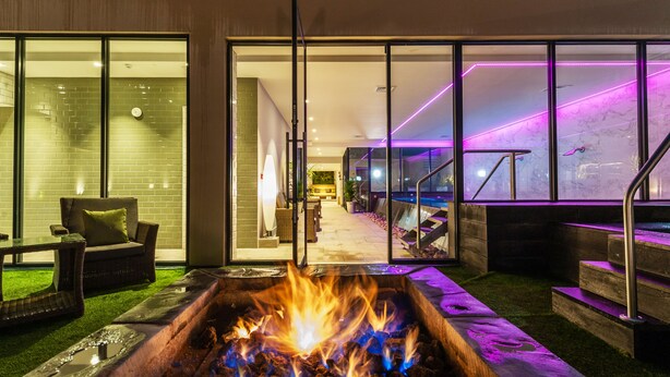 A modern indoor space with a fire pit, glass doors leading to a room with a sofa and a dining area, and a staircase with purple lighting.