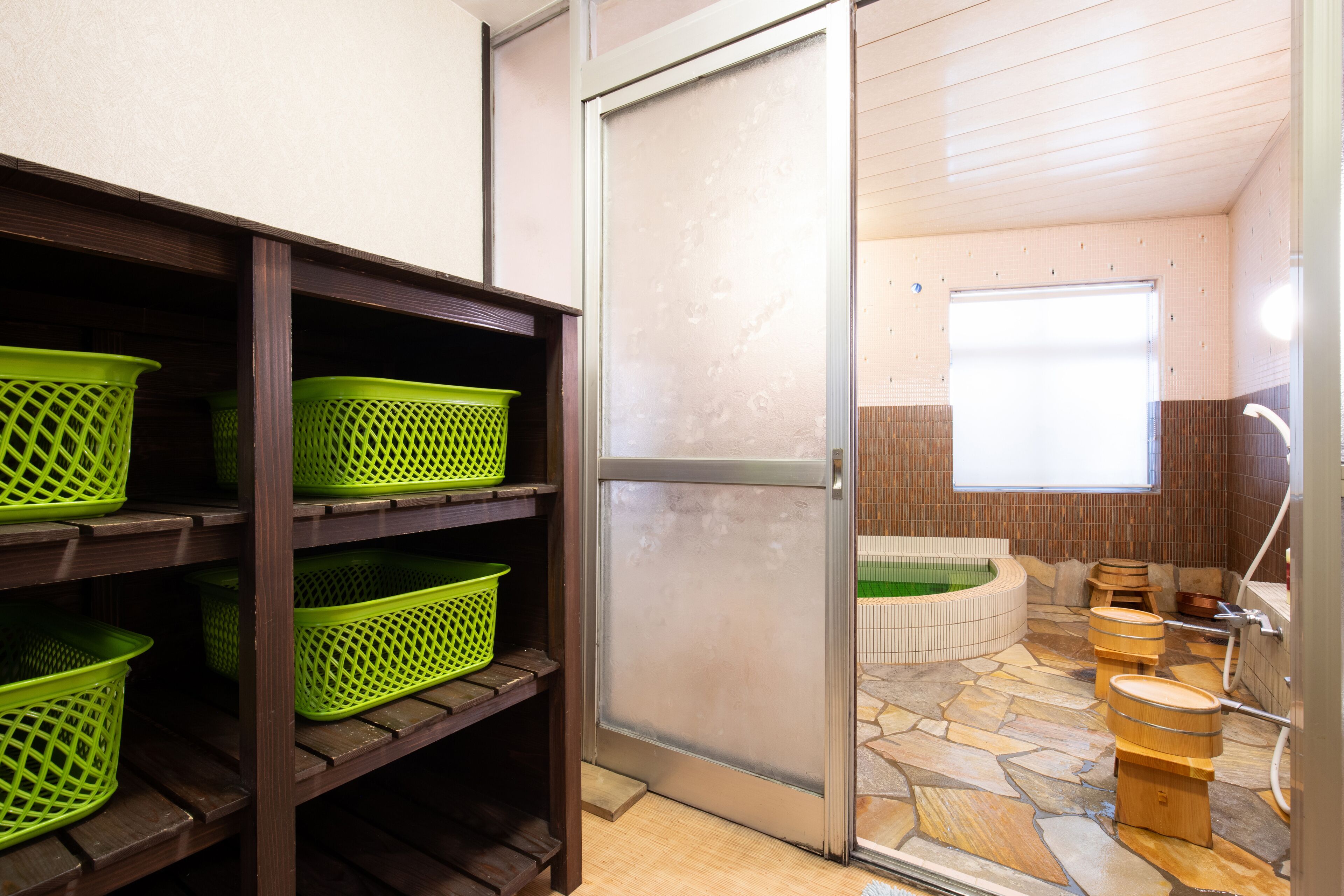A bathroom with a stone floor, wooden shelves with green baskets, a glass shower enclosure, and a bathtub.