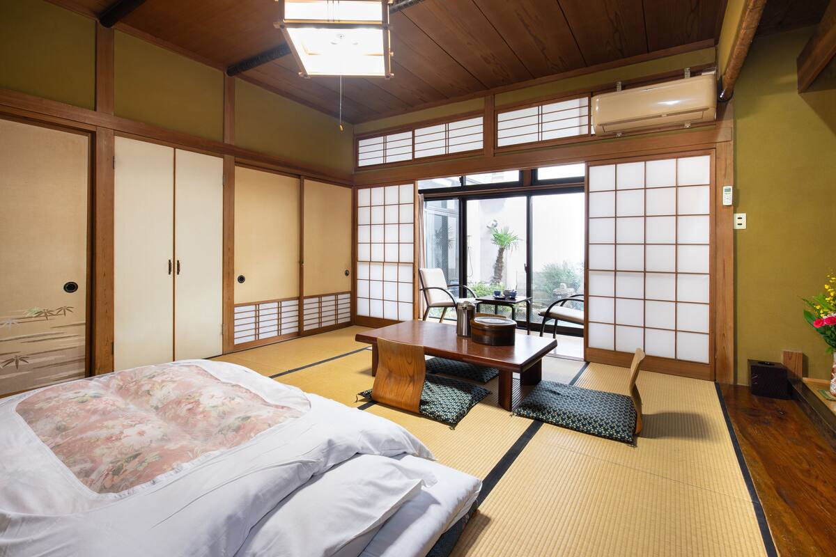 Ein traditioneller japanischer Raum mit Tatami-Böden, einem Bett mit floralem Bettbezug, einem Holztisch und Schiebetüren.