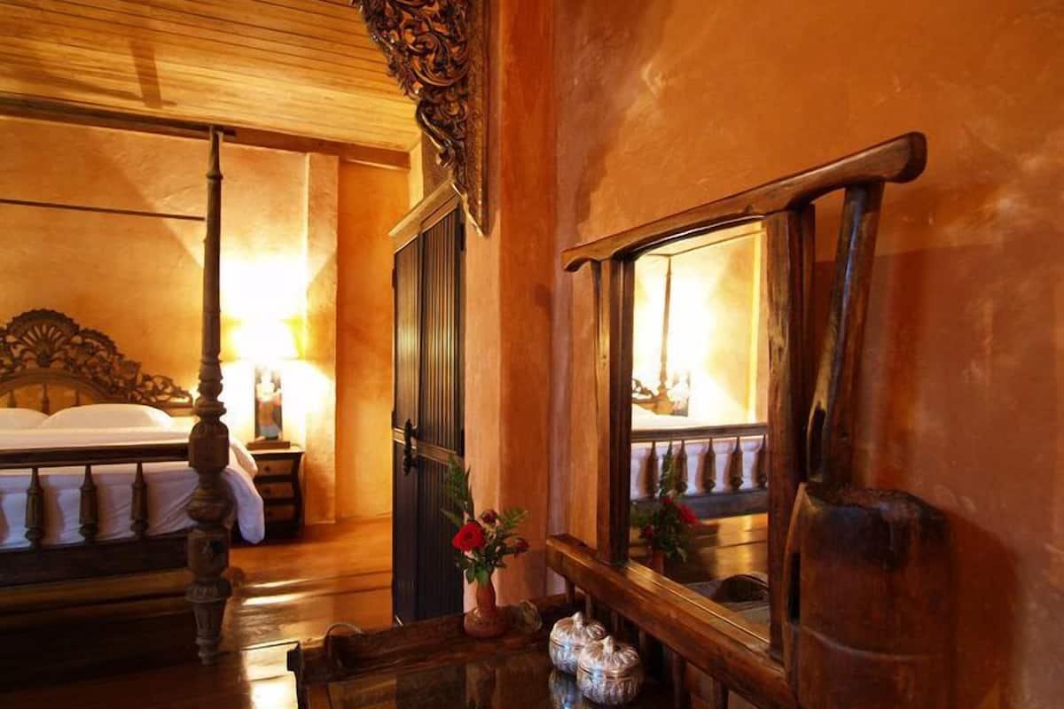 A hotel room with a four-poster bed, a nightstand with a lamp, a mirror, and a small table with decorative items.