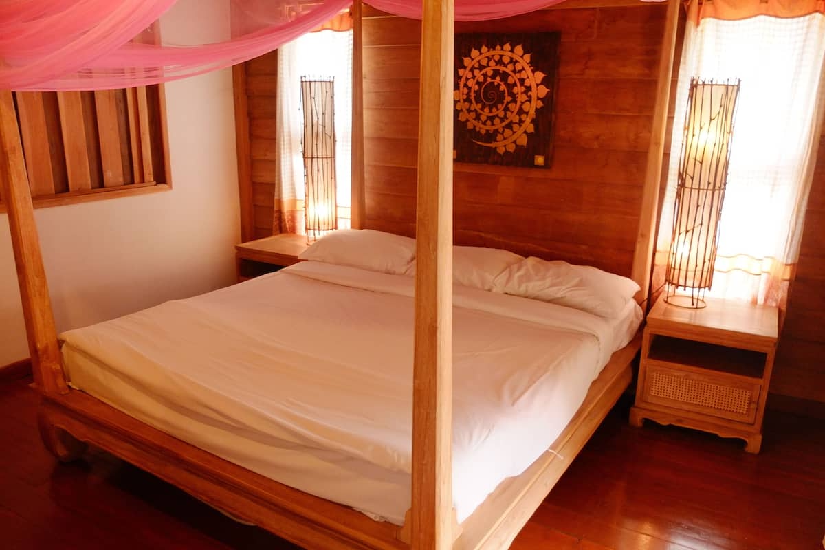A wooden four-poster bed with white linens, a wooden nightstand with a lamp, and a decorative wall piece.