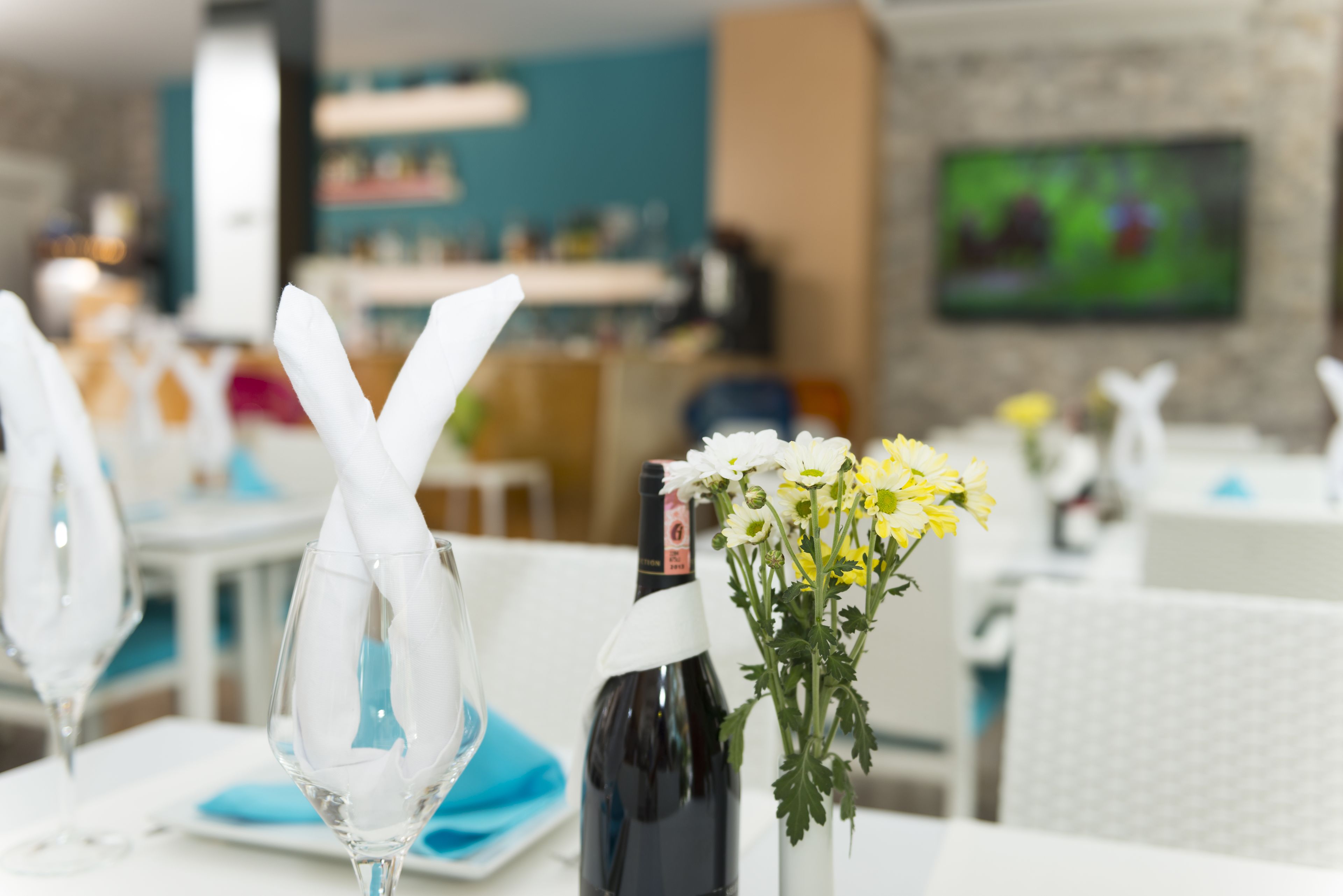 Une table à manger dressée avec une bouteille de vin, un vase rempli de fleurs jaunes et une serviette nouée en forme de nœud.