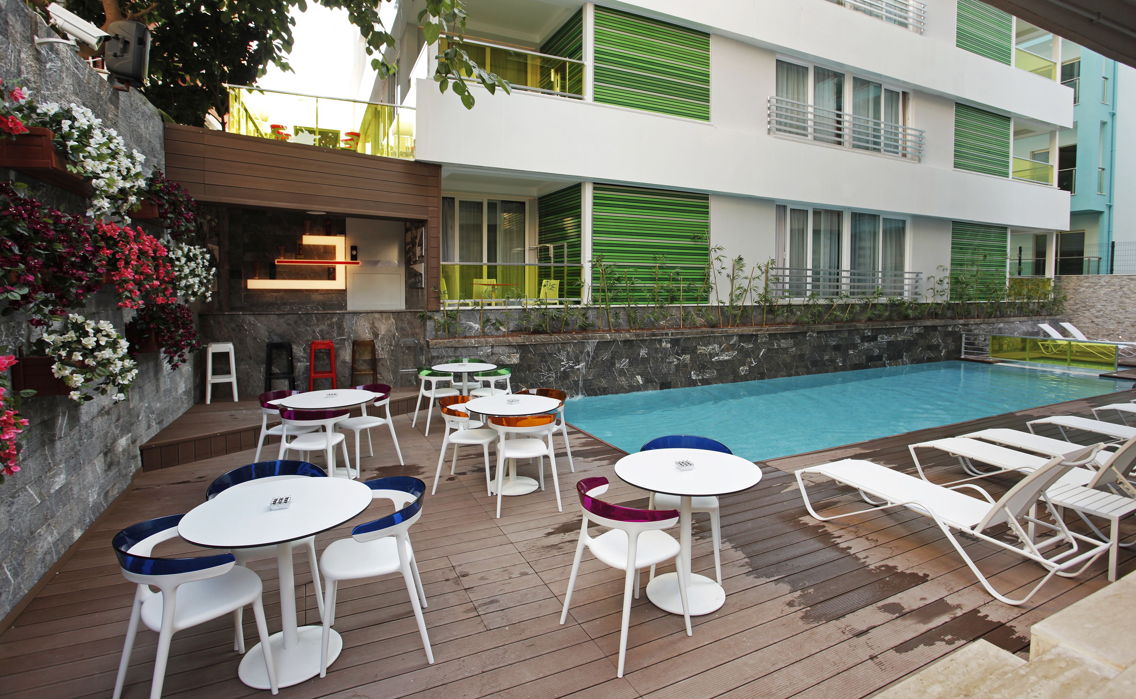 A poolside area with multiple tables and chairs, a pool, and a building with green shutters and balconies.