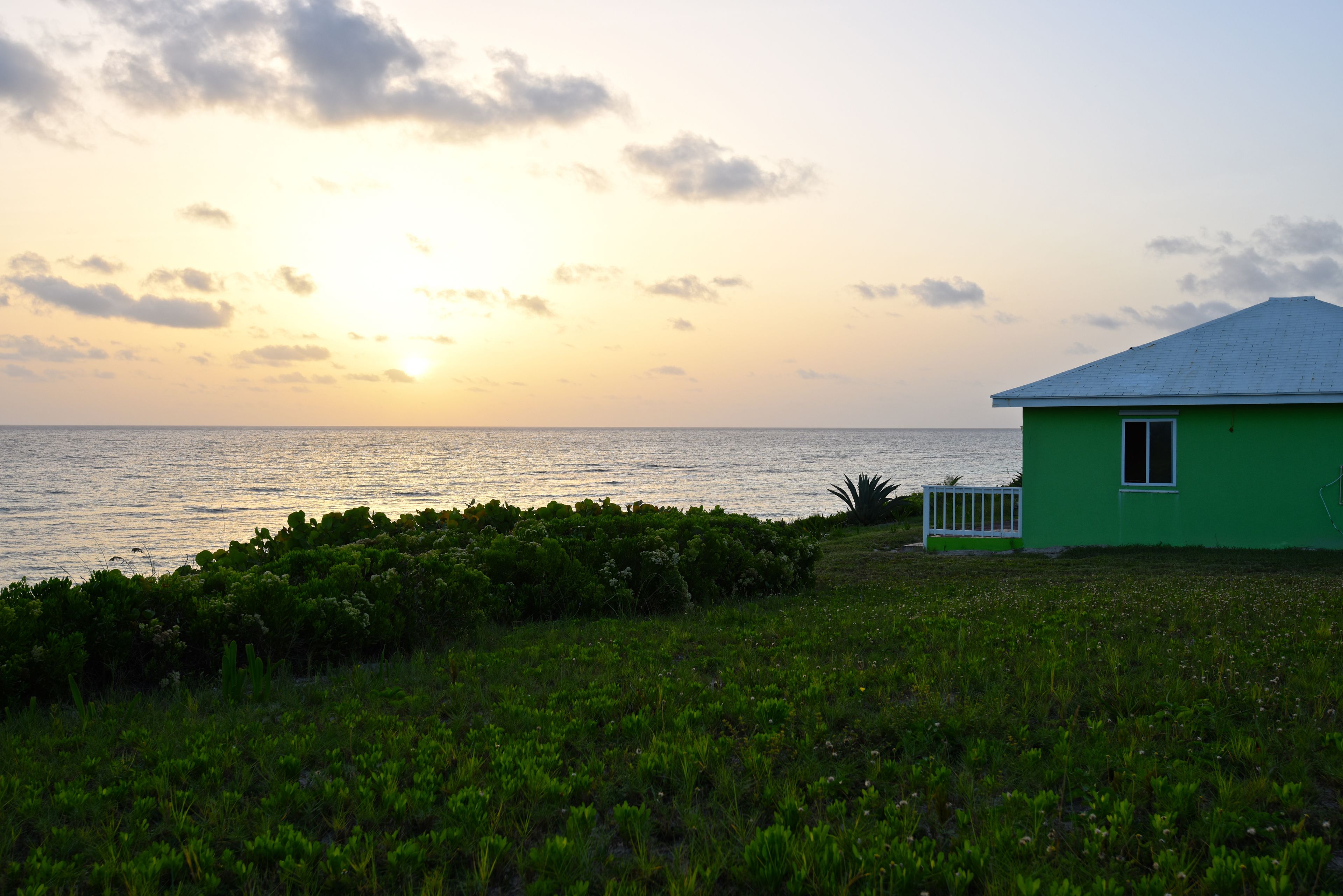 A green house with a white railing and a small garden in front, overlooking a body of water at sunset.