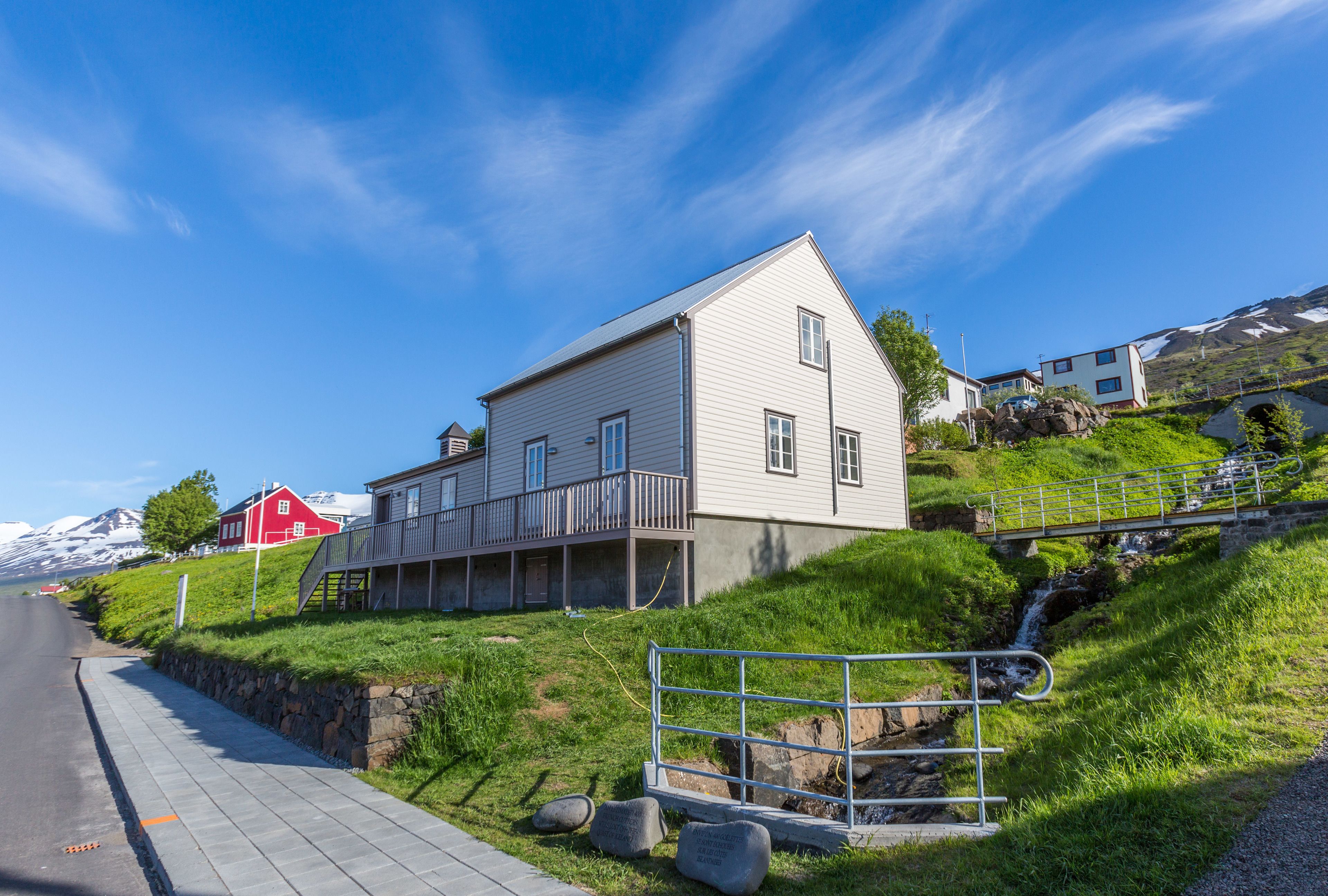 A two-story house with a balcony, located on a hillside with a view of a mountain and a road in the background.