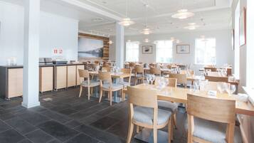 Een ruime eethoek met meerdere tafels en stoelen, een keuken op de achtergrond, en grote ramen die voor natuurlijk licht zorgen.