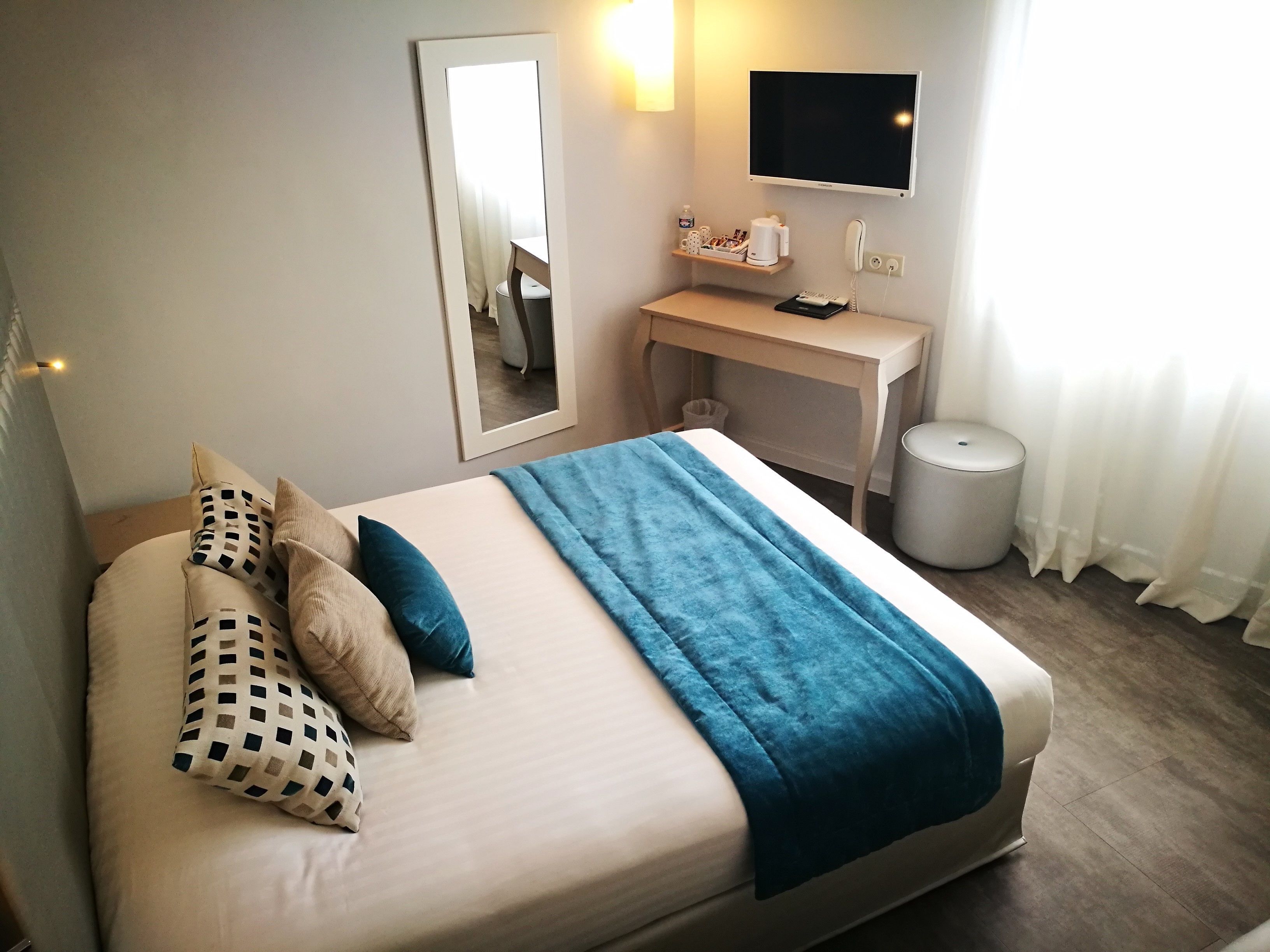 A hotel room with a bed, a desk, a chair, a mirror, a TV, and a trash bin.