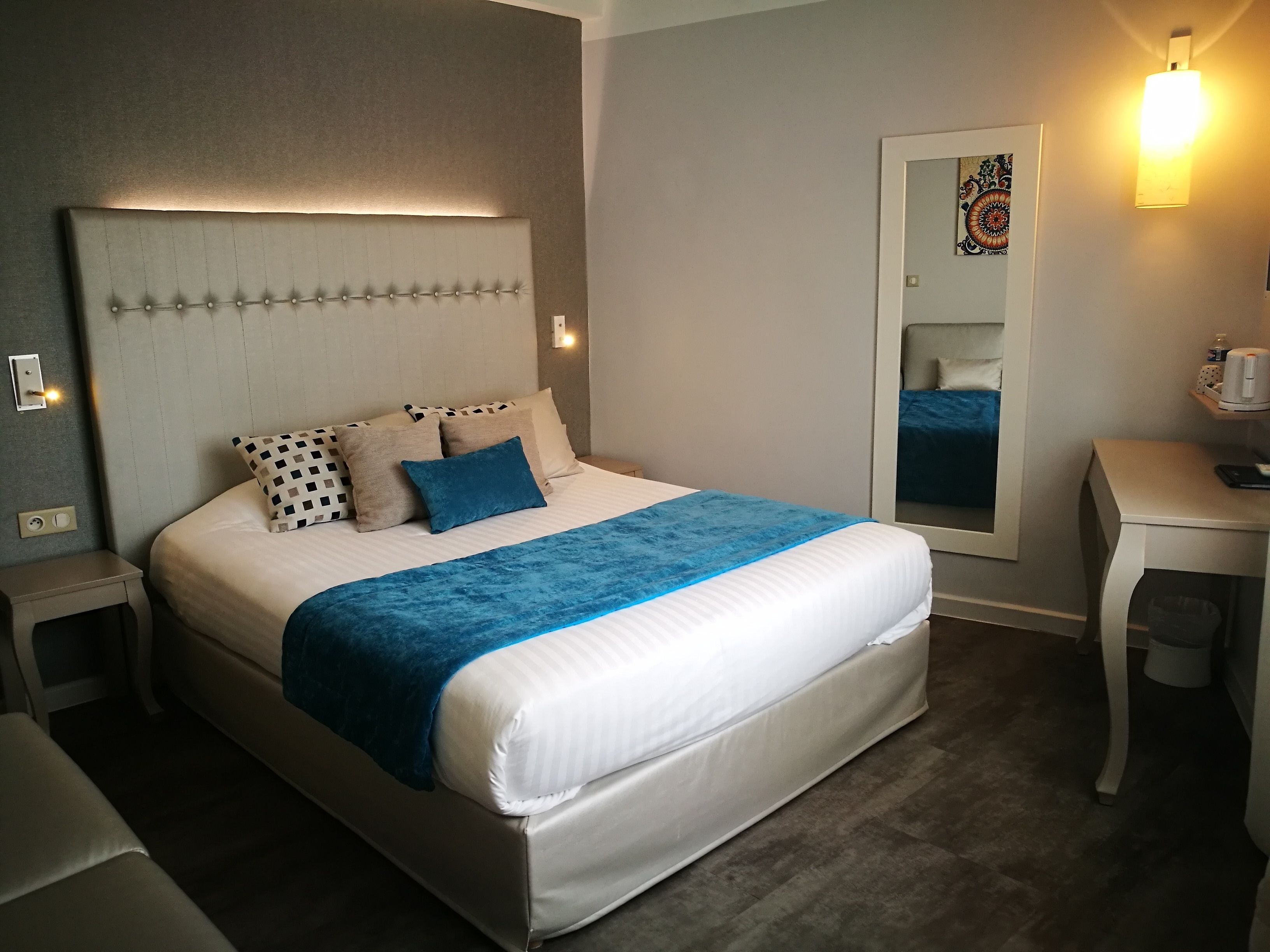 A neatly made bed with a tufted headboard, a blue throw blanket, and patterned pillows. There is a bedside table with a lamp and a small shelf with toiletries. A mirror is visible on the wall to the right.