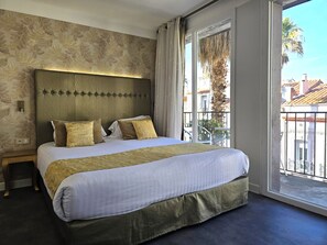 A hotel room with a large bed, a headboard with a lighted panel, a window with a view of buildings and palm trees, and a small bedside table.