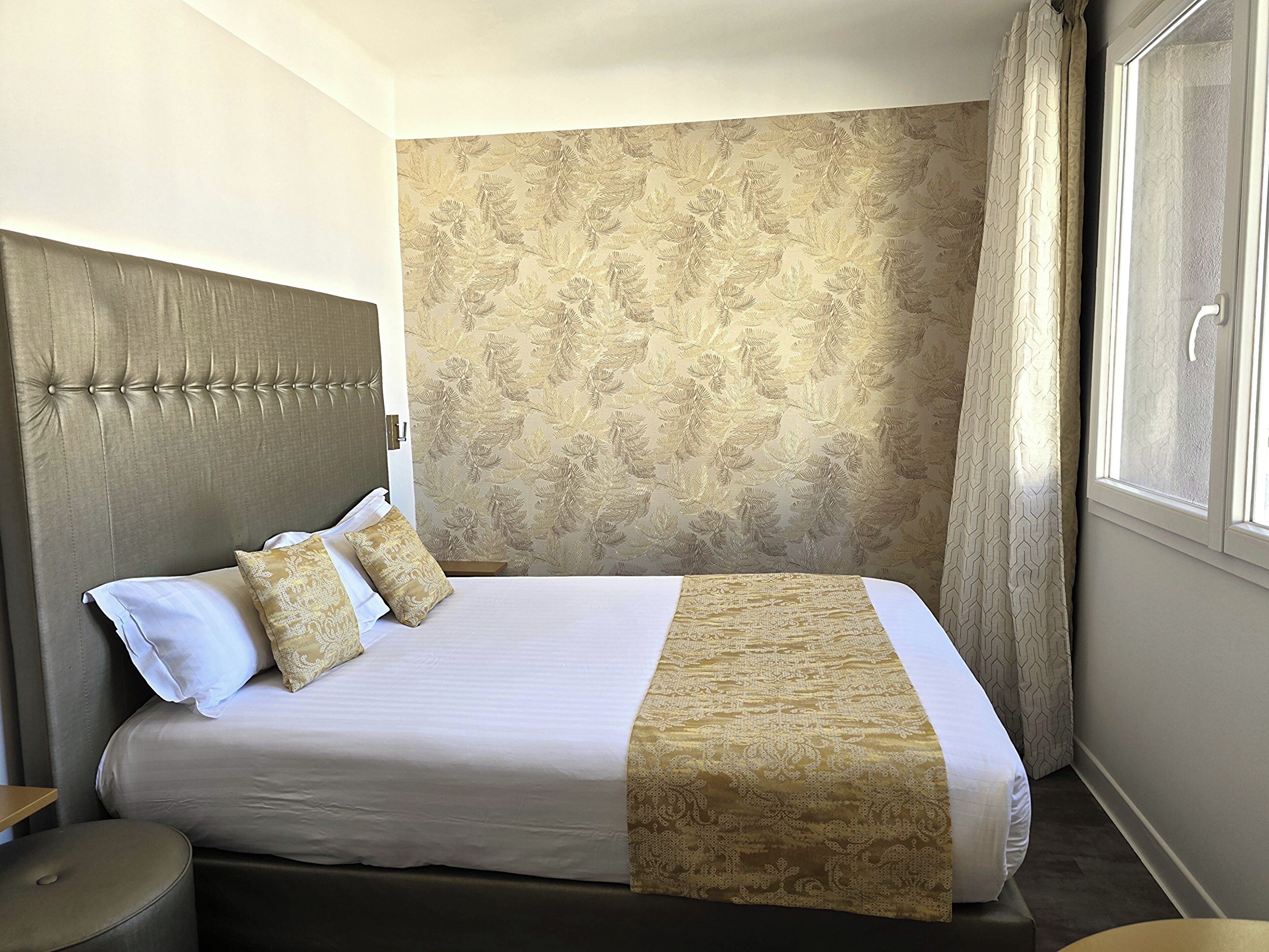 Un lit bien fait, avec une couvre-lit à motifs et deux oreillers, une tête de lit capitonnée et une fenêtre avec des rideaux.