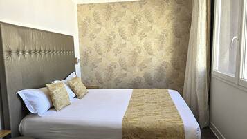 Un lit bien fait, avec une couvre-lit à motifs et deux oreillers, une tête de lit capitonnée et une fenêtre avec des rideaux.
