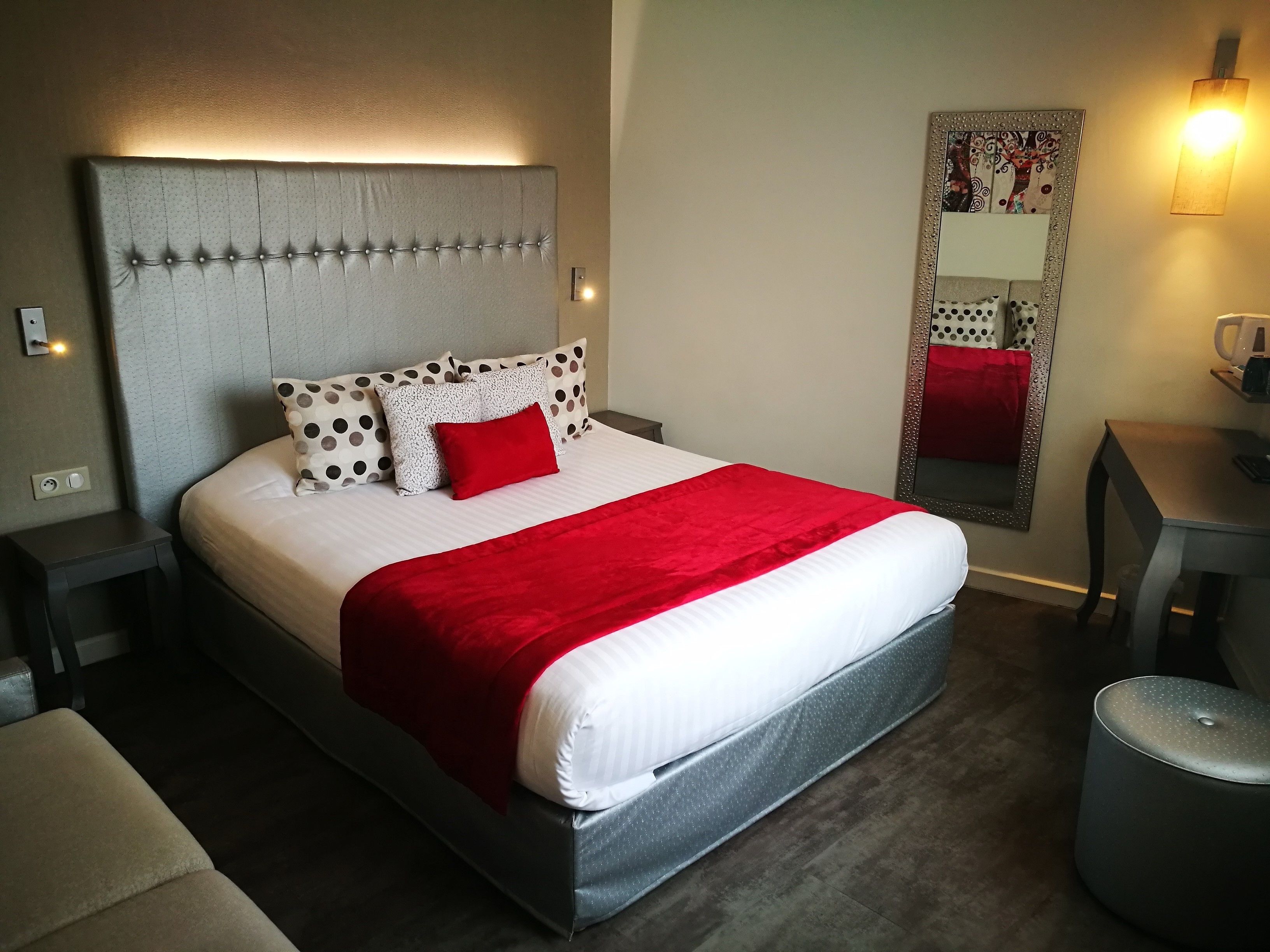 A neatly arranged hotel room with a large bed, a bedside table, a chair, a mirror, and a wall-mounted lamp.