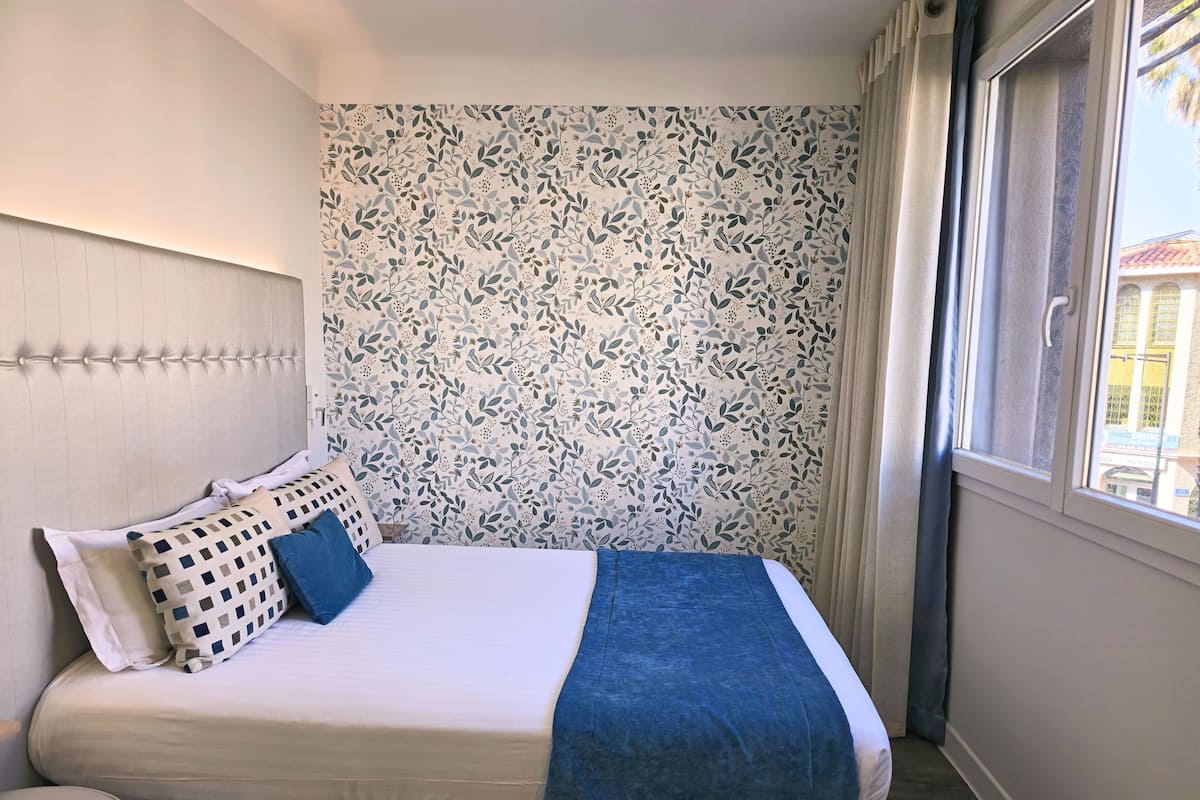 Hotellihuone, jossa on sänky, sängynpääty, ikkuna verhoilla ja näkymä ulkona oleviin rakennuksiin.
