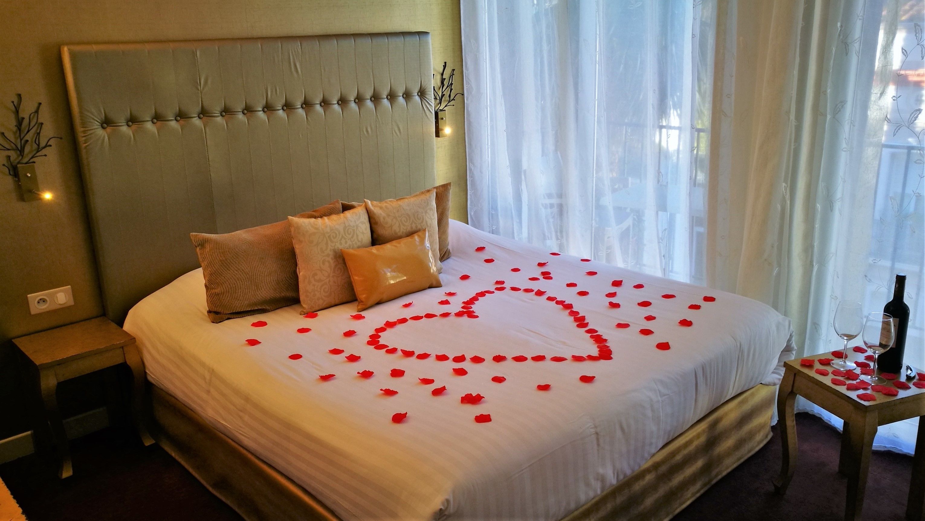 A neatly made bed with rose petals arranged in a heart shape, a bottle of wine, and a glass on a small table beside the bed.