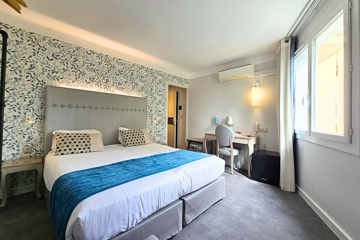 Hotellihuoneessa on suuri sänky, työpöytä ja tuoli. Huoneessa on ikkuna, jossa on verhot, seinälle asennettu ilmastointilaite ja ovi.