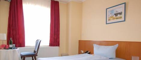 Habitación de hotel con una cama, una silla, una mesa con una botella y un cuadro en la pared.