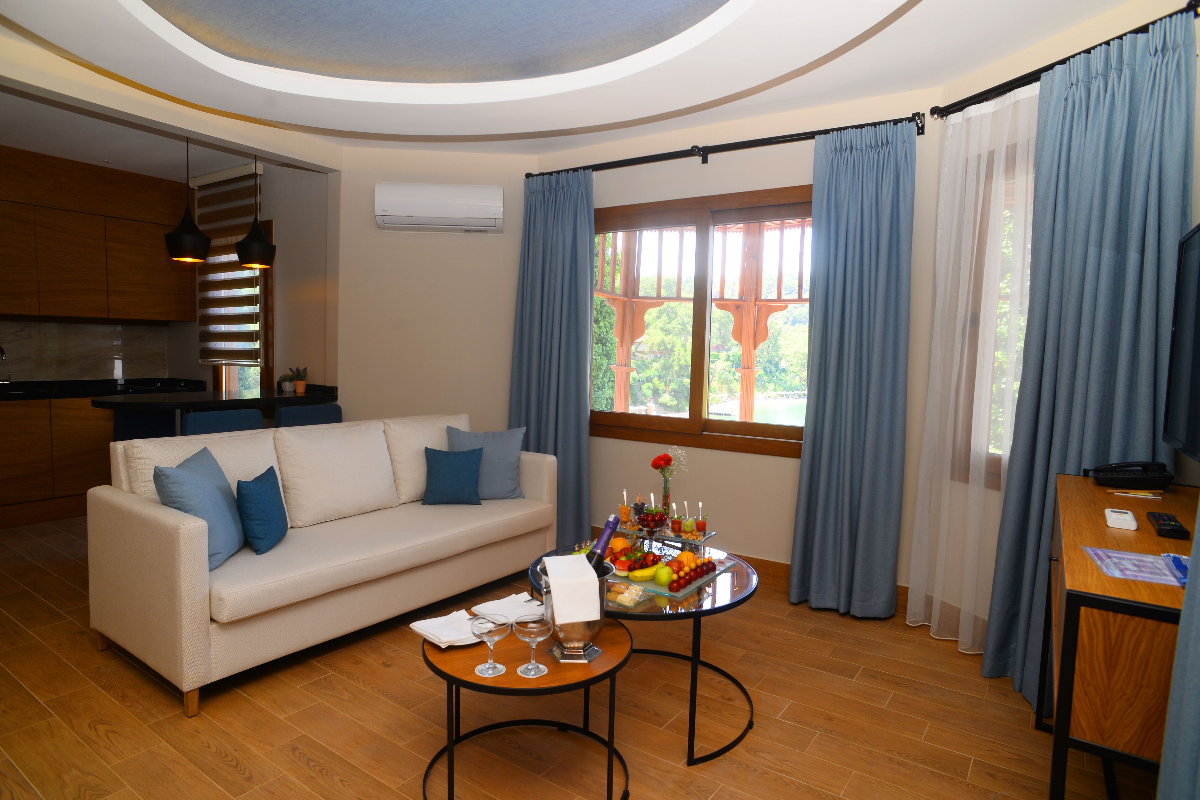 A modern living room with a sofa, a coffee table with drinks and snacks, a kitchen area, and large windows with blue curtains.