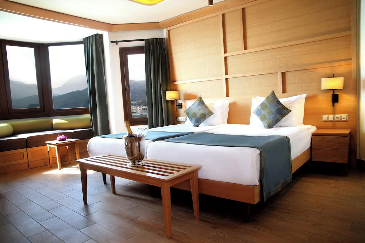 A hotel room with a large bed, a wooden nightstand, a small table, and a view of mountains through the window.
