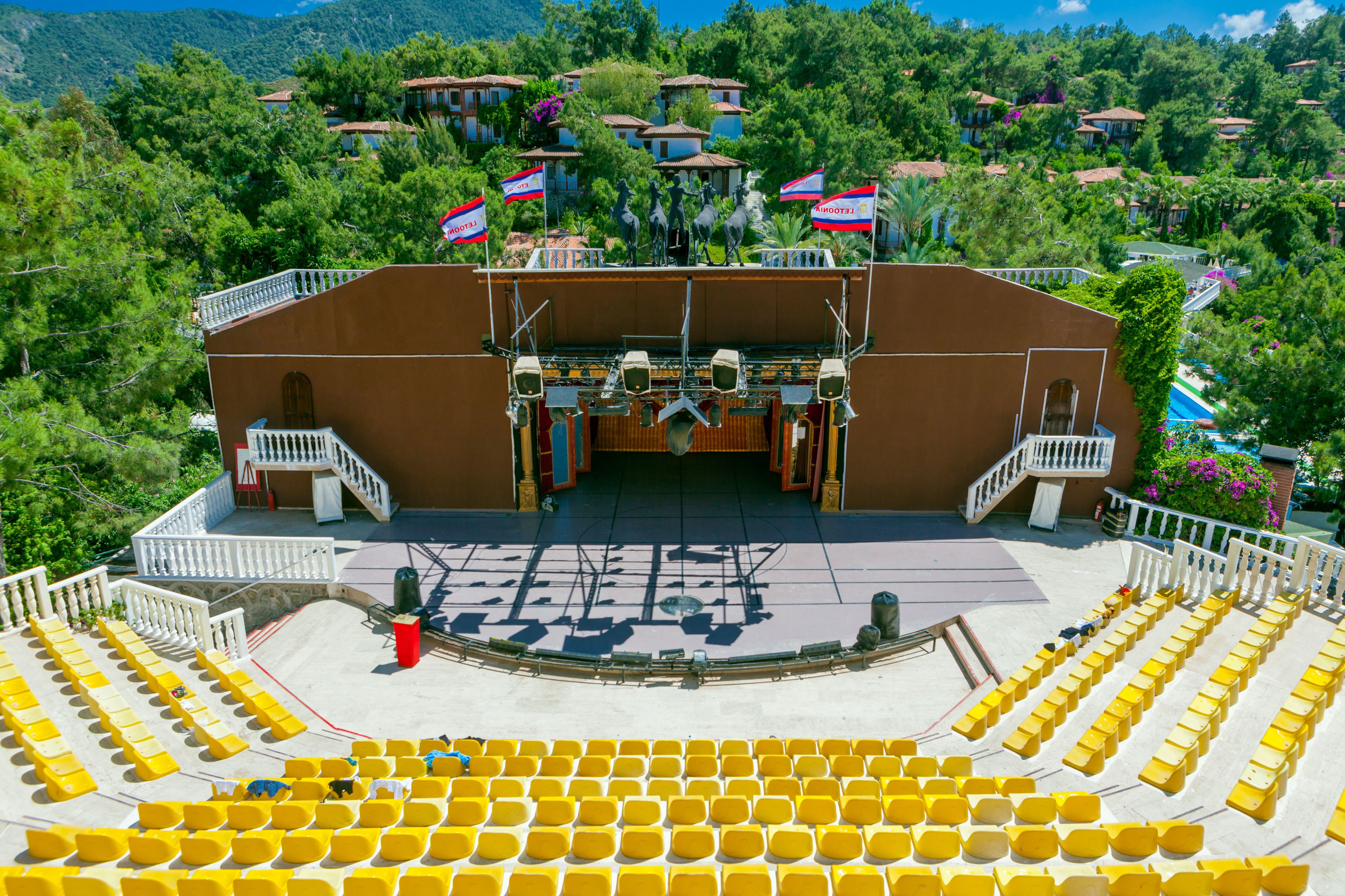An outdoor amphitheater with tiered seating and a stage, surrounded by trees and buildings.