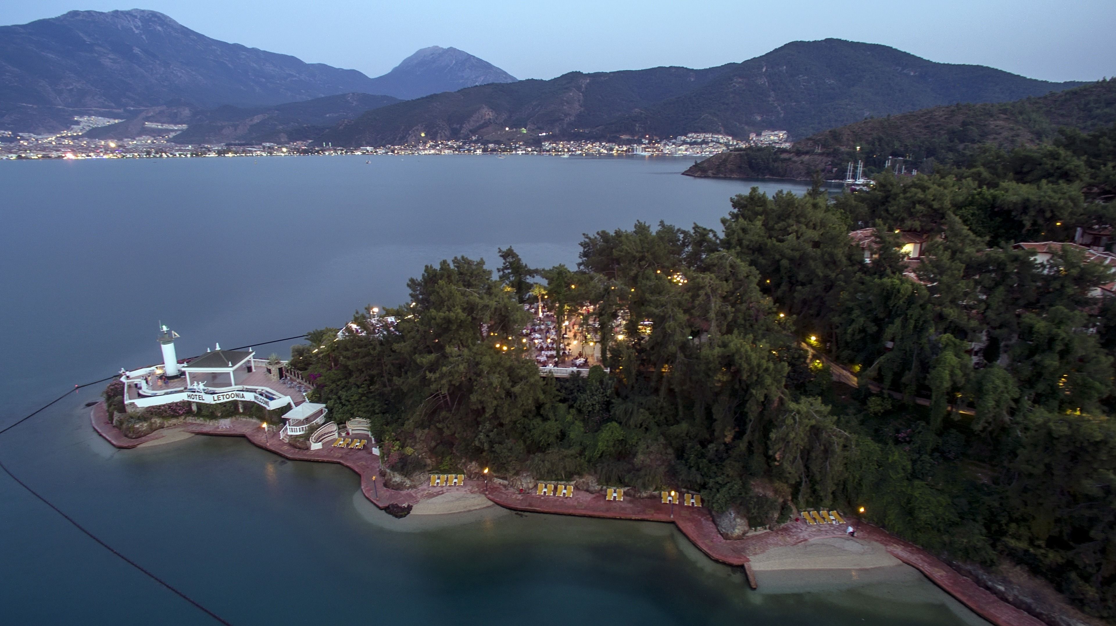 A coastal hotel with a lighthouse, surrounded by dense forest and mountains.