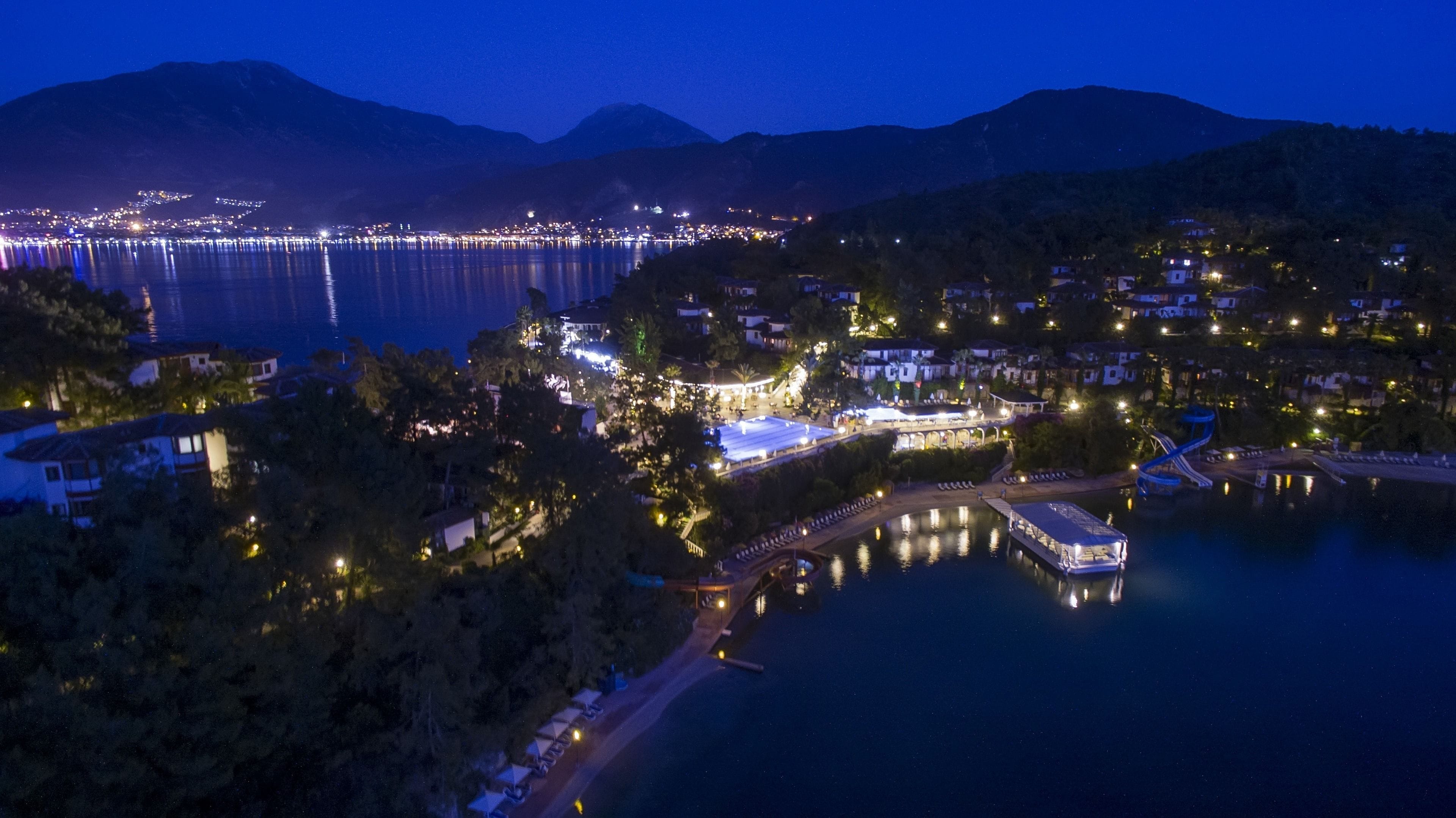 A coastal town at night with a marina, illuminated buildings, and a mountain backdrop.