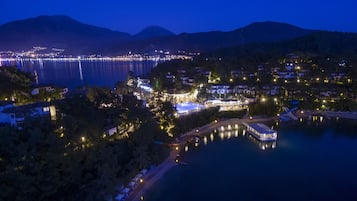 A coastal town at night with a marina, illuminated buildings, and a mountain backdrop.